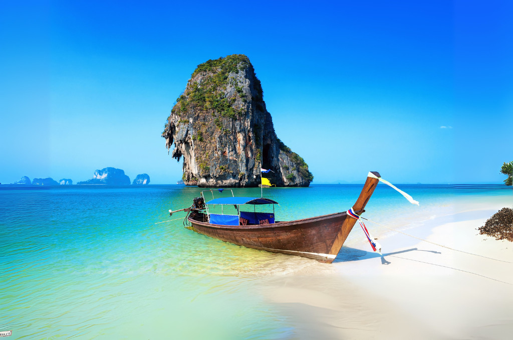 Thailändisches Longtail-Boot an einem weißen Strand vor einem riesigen, bewachsenen Kalksteinfelsen im klaren, türkisen Meer unter blauem Himmel. Thailändisches Longtail-Boot an einem weißen Strand vor einem riesigen, bewachsenen Kalksteinfelsen im klaren, türkisen Meer unter blauem Himmel.