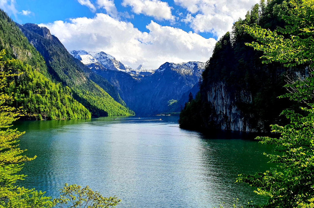 Blick auf einen ruhigen, grünen See, umgeben von steilen, bewaldeten Bergen. Im Hintergrund sind schneebedeckte Gipfel sichtbar. Der Himmel ist blau mit weißen Wolken, und das Wasser spiegelt teilweise die Landschaft wider. Im Vordergrund rahmen grüne Bäume den See ein.