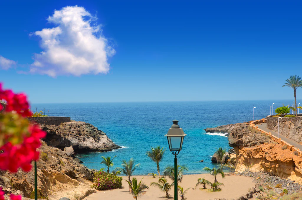 Eine tropische Meeresbucht mit feinem Sandstrand und Palmen unter einem strahlend blauen Himmel mit einer einzelnen weißen Wolke. Im Vordergrund sind eine klassische Straßenlaterne sowie leuchtend rote Blüten im Anschnitt zu sehen; raue Klippen umrahmen das türkisfarbene Wasser.