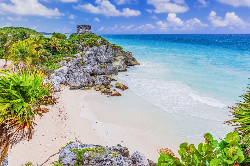 Blick auf die Maya-Ruinen von Tulum auf einer Klippe über einem weißen Sandstrand und türkisblauen Meer. Im Vordergrund Palmen und Felsen.