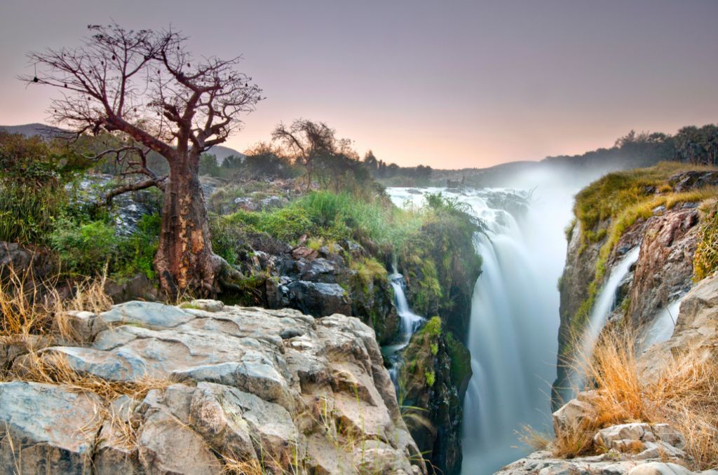 Epupa-Wasserfälle in Namibia: Ein mächtiger Wasserfall stürzt in eine Schlucht, daneben ein markanter Affenbrotbaum (Baobab) auf Felsen im Abendlicht.