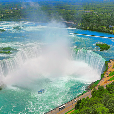 Ein hoher Blickwinkel auf die Hufeisenfälle der Niagarafälle, die massiv sind und eine riesige Menge Wasser über eine weite hufeisenförmige Klippe in ein türkisfarbenes Becken darunter stürzen lassen. Die Gischt von den Fällen steigt hoch in die Luft auf und schafft eine nebelige Atmosphäre. Im Becken darunter fährt ein kleines weiß-blaues Touristenboot durch das raue Wasser und scheint in Richtung des Wasserfalls zu fahren. Ein breiter Bürgersteig am rechten unteren Bildrand ist voller Menschen, die die Fälle beobachten, und eine Straße verläuft parallel dazu, auf der ein weißer Bus fährt. Im Hintergrund ist die breite Niagarafälle in weitere grüne Wälder und ferne Gebäude eingebettet. Ein hoher Blickwinkel auf die Hufeisenfälle der Niagarafälle, die massiv sind und eine riesige Menge Wasser über eine weite hufeisenförmige Klippe in ein türkisfarbenes Becken darunter stürzen lassen. Die Gischt von den Fällen steigt hoch in die Luft auf und schafft eine nebelige Atmosphäre. Im Becken darunter fährt ein kleines weiß-blaues Touristenboot durch das raue Wasser und scheint in Richtung des Wasserfalls zu fahren. Ein breiter Bürgersteig am rechten unteren Bildrand ist voller Menschen, die die Fälle beobachten, und eine Straße verläuft parallel dazu, auf der ein weißer Bus fährt. Im Hintergrund ist die breite Niagarafälle in weitere grüne Wälder und ferne Gebäude eingebettet.