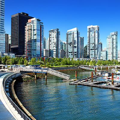 Ein Stadtbild von Vancouver, Kanada, mit einem Yachthafen im Vordergrund und hohen Gebäuden im Hintergrund. Das Wasser im Yachthafen ist dunkelblau, mit vielen leeren Bootsstegen, die ins Bild ragen. Am linken Rand des Bildes verläuft ein breiter, gebogener Weg entlang des Wassers. Dahinter, auf der Uferpromenade, sind grüne Bäume und niedrige Büsche zu sehen, gefolgt von einer Reihe moderner Hochhäuser aus Glas und Stahl, die in den klaren, hellblauen Himmel ragen. Ein einzelnes dunkles Gebäude sticht inmitten der hellen Gebäude hervor. Ein Stadtbild von Vancouver, Kanada, mit einem Yachthafen im Vordergrund und hohen Gebäuden im Hintergrund. Das Wasser im Yachthafen ist dunkelblau, mit vielen leeren Bootsstegen, die ins Bild ragen. Am linken Rand des Bildes verläuft ein breiter, gebogener Weg entlang des Wassers. Dahinter, auf der Uferpromenade, sind grüne Bäume und niedrige Büsche zu sehen, gefolgt von einer Reihe moderner Hochhäuser aus Glas und Stahl, die in den klaren, hellblauen Himmel ragen. Ein einzelnes dunkles Gebäude sticht inmitten der hellen Gebäude hervor.