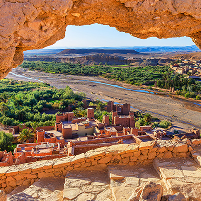 Ein Panoramablick auf die Lehmstadt Aït-Ben-Haddou in Marokko, die durch eine natürliche Felsformation im Vordergrund eingerahmt ist. Die Stadt besteht aus beigefarbenen Lehmgebäuden, die sich auf einem Hügel scharen, und ist von Palmen umgeben. Ein Flussbett, das teilweise Wasser führt, schlängelt sich durch ein grünes Tal im Hintergrund, gefolgt von kahlen, braunen Bergen unter einem klaren blauen Himmel. Der Felsrahmen im Vordergrund ist bräunlich-orange und rau. Ein Panoramablick auf die Lehmstadt Aït-Ben-Haddou in Marokko, die durch eine natürliche Felsformation im Vordergrund eingerahmt ist. Die Stadt besteht aus beigefarbenen Lehmgebäuden, die sich auf einem Hügel scharen, und ist von Palmen umgeben. Ein Flussbett, das teilweise Wasser führt, schlängelt sich durch ein grünes Tal im Hintergrund, gefolgt von kahlen, braunen Bergen unter einem klaren blauen Himmel. Der Felsrahmen im Vordergrund ist bräunlich-orange und rau.