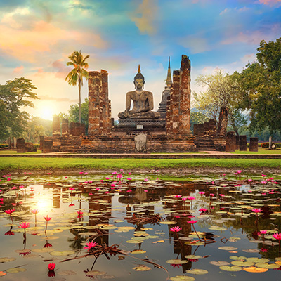 Ein großes steinernes Buddha-Bildnis sitzt auf einer erhöhten Plattform inmitten der Ruinen eines alten Tempels mit zerfallenen Säulen. Im Vordergrund befindet sich ein Teich, der mit Seerosen und rosa Lotusblüten bedeckt ist und die Spiegelung des Himmels und des Tempels zeigt. Im Hintergrund ragen Palmen und andere Bäume in den Himmel, der von den Farben des Sonnenuntergangs - Orange, Rosa, Gelb und Blau - gezeichnet ist. Die Sonne scheint von links hinter den Bäumen hervor. Ein großes steinernes Buddha-Bildnis sitzt auf einer erhöhten Plattform inmitten der Ruinen eines alten Tempels mit zerfallenen Säulen. Im Vordergrund befindet sich ein Teich, der mit Seerosen und rosa Lotusblüten bedeckt ist und die Spiegelung des Himmels und des Tempels zeigt. Im Hintergrund ragen Palmen und andere Bäume in den Himmel, der von den Farben des Sonnenuntergangs - Orange, Rosa, Gelb und Blau - gezeichnet ist. Die Sonne scheint von links hinter den Bäumen hervor.