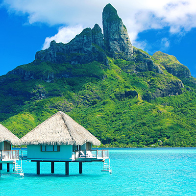 Ein Bild von Overwater-Bungalows mit Strohdächern in einem türkisfarbenen Meer vor einem großen, üppig grünen Berg unter einem blauen Himmel mit einigen Wolken. Die Bungalows stehen auf Stelzen im Wasser, und Stufen führen direkt ins Meer. Ein Bild von Overwater-Bungalows mit Strohdächern in einem türkisfarbenen Meer vor einem großen, üppig grünen Berg unter einem blauen Himmel mit einigen Wolken. Die Bungalows stehen auf Stelzen im Wasser, und Stufen führen direkt ins Meer.
